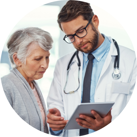 A doctor in a white coat with a stethoscope holds a tablet and discusses something with an elderly woman in a clinical setting.