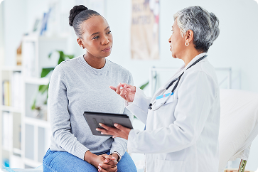 Two people seated in a clinical office, a doctor holding a tablet and speaking while the other listens.