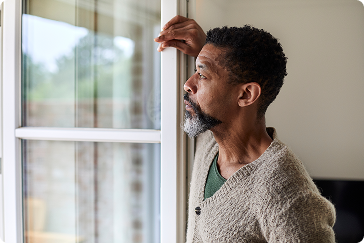 Person standing indoors by a window, looking outside with one hand resting on the window frame..