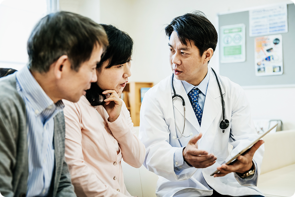 A doctor shows a tablet while speaking with two seated people in a medical office.