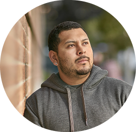 Close-up portrait of a person wearing a gray hoodie, standing beside a brick wall and looking slightly upward outdoors, with a softly blurred background