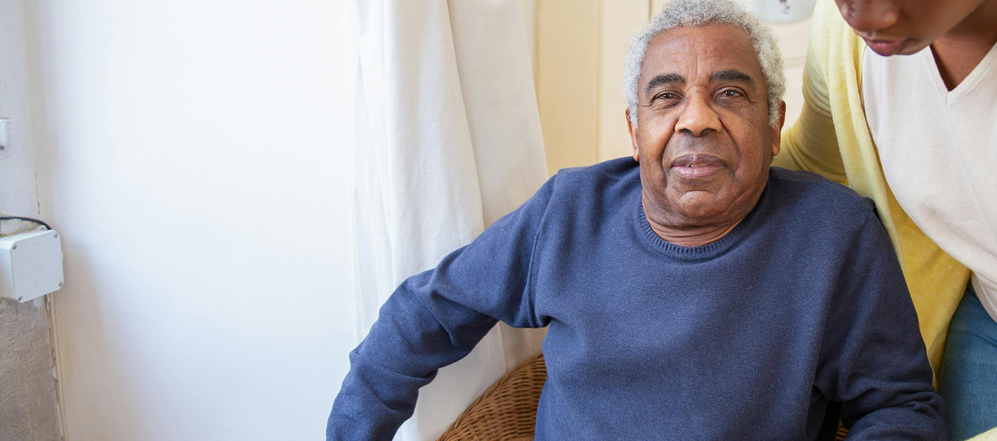 An elderly male between 60-70 years with gray hair, wearing a blue sweater, is sitting down. Another individual, wearing a white shirt and yellow cardigan, is standing beside the elderly person and providing support by placing a hand on the elderly person's shoulder.