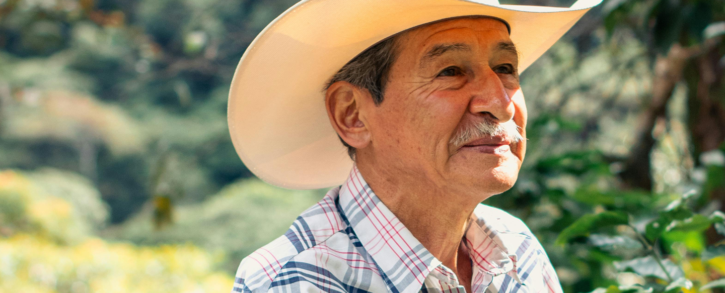 An elderly male wearing a cowboy hat and a plaid shirt. The background appears to be outdoors with greenery visible.